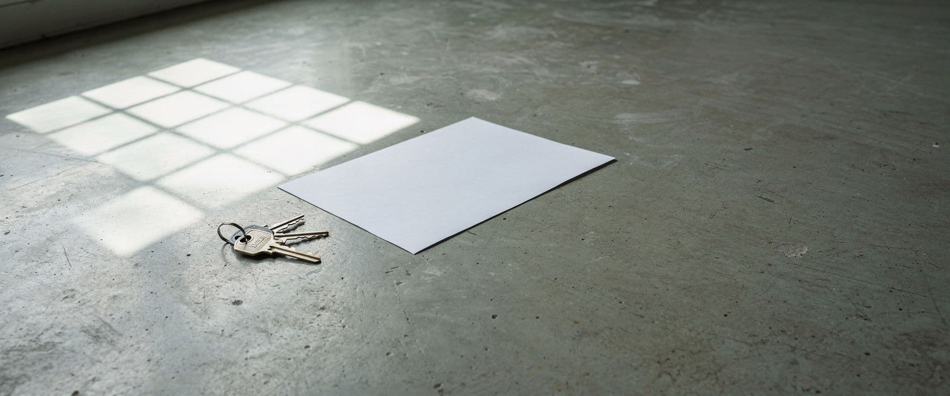 Keys and a blank sheet of paper on the bare floor of an empty flat, lit by a grid of window light.